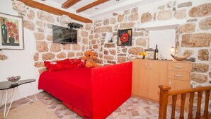 Une pièce avec un mur en pierre, un lit rouge, une armoire en bois et un téléviseur fixé au mur.