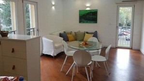 A dining area with a glass table, white chairs, a sofa with cushions, and a painting on the wall. - Relaxing Tivat Stay with Balcony -Two Bedroom Apt (Tivat)
