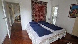 A bedroom with a bed on a white pallet base, a patterned tapestry on the wall, and a window with blinds. - Relaxing Tivat Stay with Balcony -Two Bedroom Apt (Tivat)
