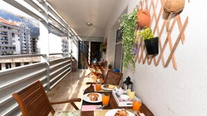 A balcony with wooden chairs and a table set for breakfast, overlooking a cityscape.