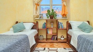 Una habitación con dos camas, una ventana con cortinas, una estantería y vistas a edificios.