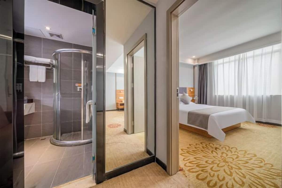 A hotel room with a glass-enclosed shower, a bed with a white bedspread, and a large window with curtains.