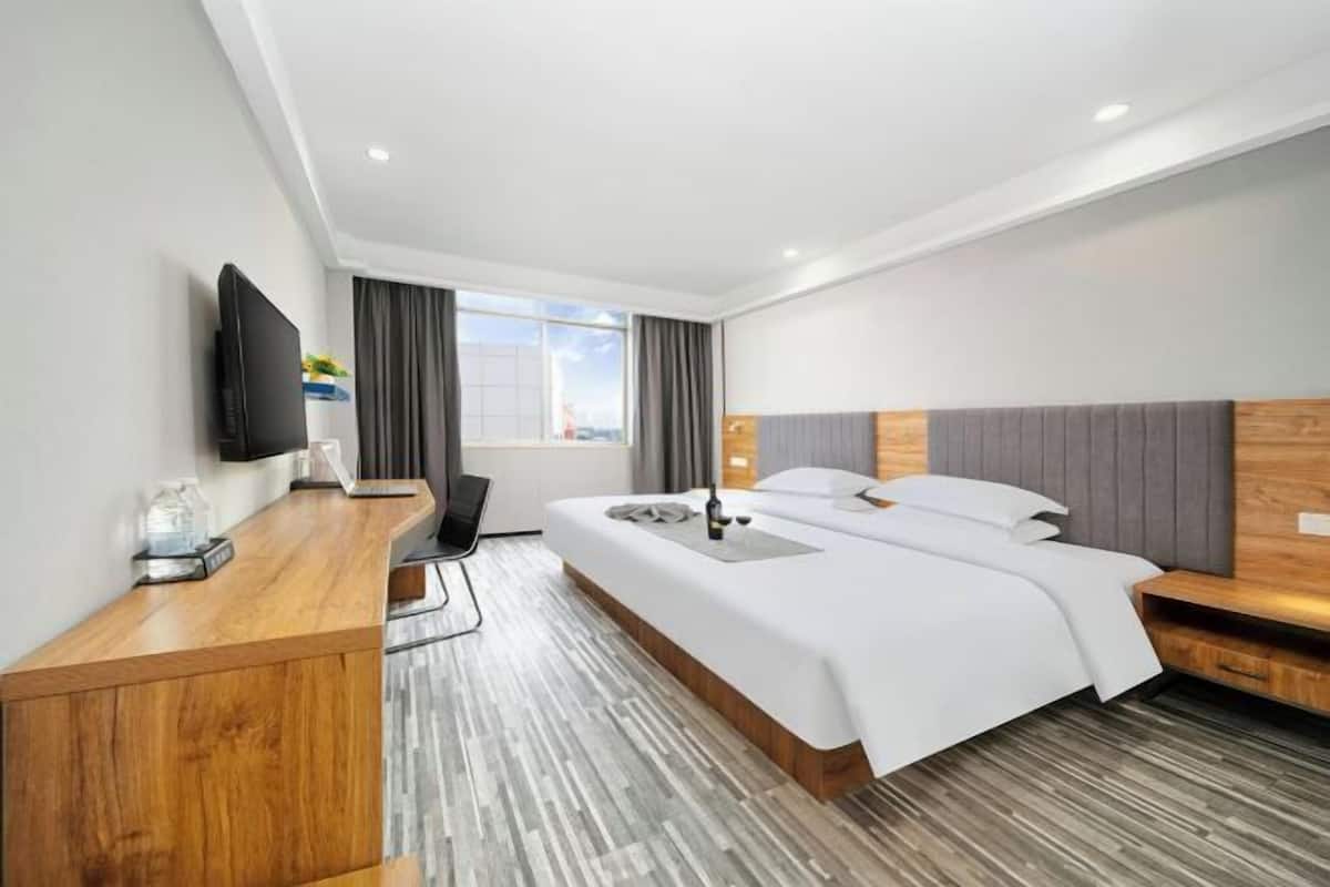 A modern hotel room with a wooden desk, a bed with a grey headboard, a flat-screen TV, and a large window.