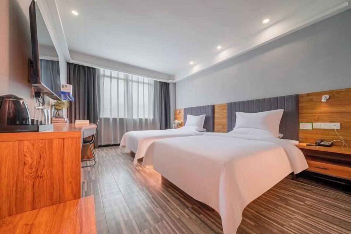 A hotel room with two beds, a wooden desk, a flat-screen TV, and a large window with curtains.