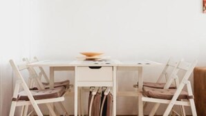 Un coin repas avec une table et des chaises blanches, un bol posé sur la table et un mur blanc orné d’un tableau encadré.