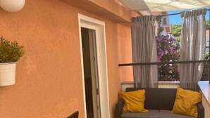 Ein Balkon mit einer Couch und Kissen, einer Topfpflanze und Blick auf ein Gebäude und Bäume.