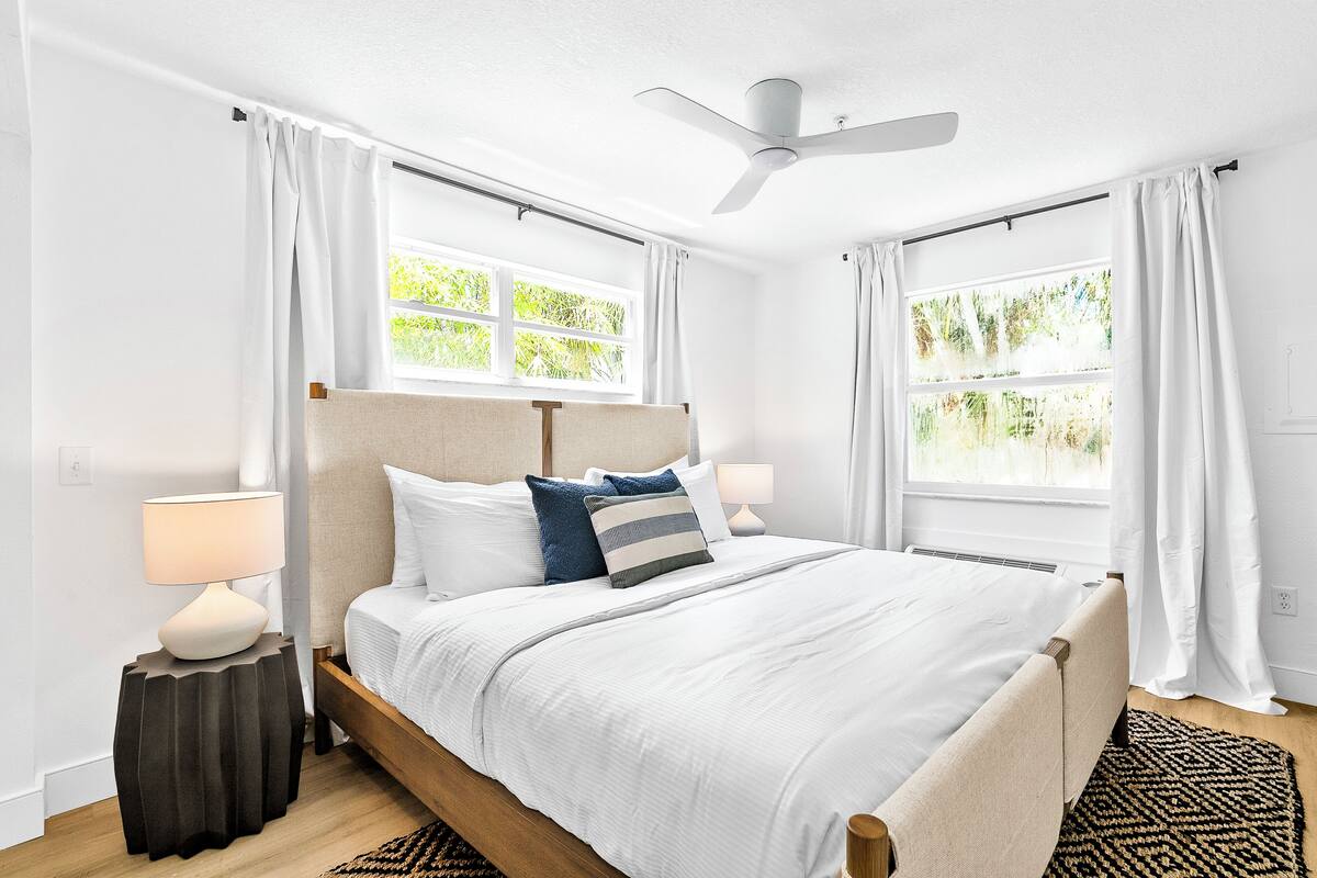A bedroom with a bed, two bedside tables, a ceiling fan, and windows with curtains.