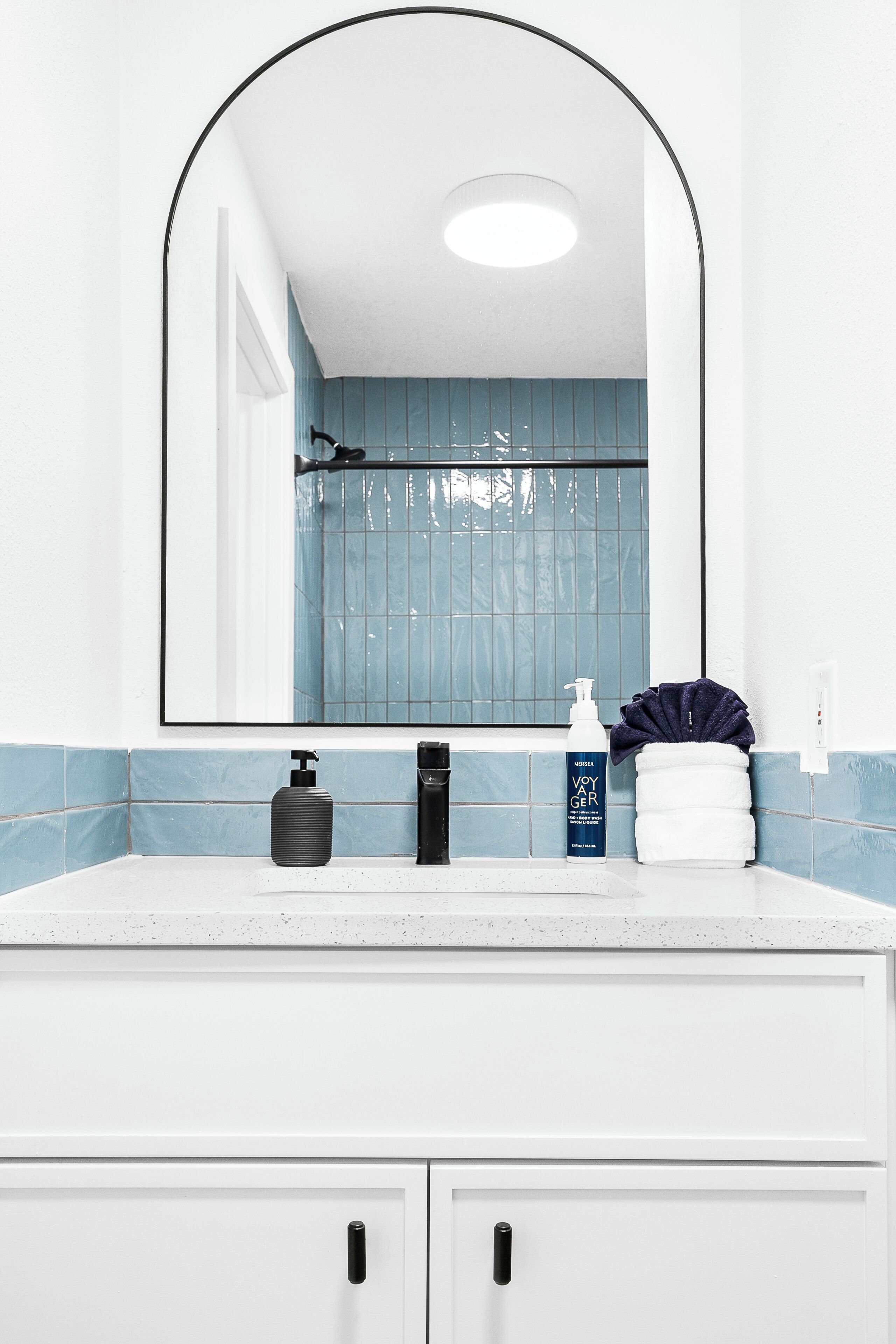 A bathroom with a white vanity, a mirror with an arch frame, a blue-tiled shower area, and toiletries on the counter.