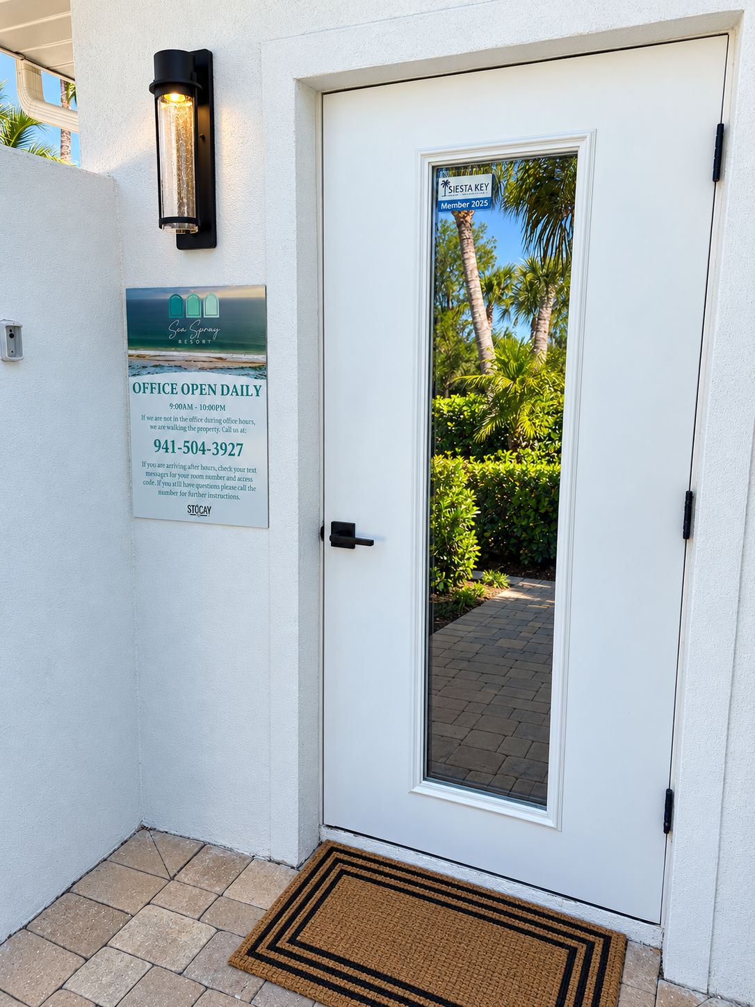 A white door with a glass panel, a doormat, a wall-mounted light, and a sign indicating office hours and a contact number.