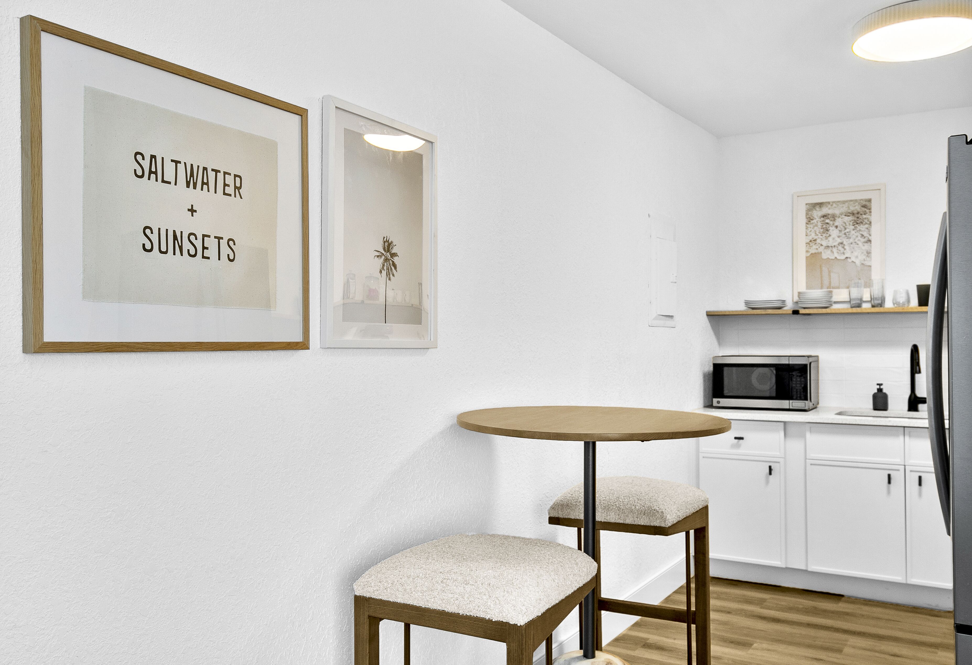 Une cuisine moderne avec des armoires blanches, un four à micro-ondes et une table ronde accompagnée de deux chaises. Des tableaux encadrés ornent le mur.