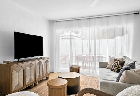 A modern living room with a flat-screen TV on a wooden cabinet, a grey sofa with patterned cushions, and a large window with sheer curtains.