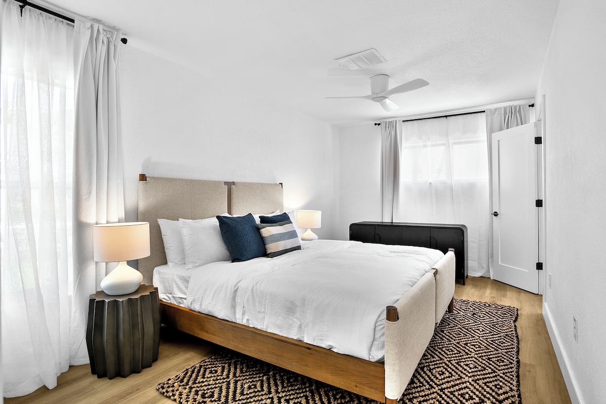 A modern bedroom with a wooden bed, white bedding, a side table with a lamp, a patterned rug, and a ceiling fan.