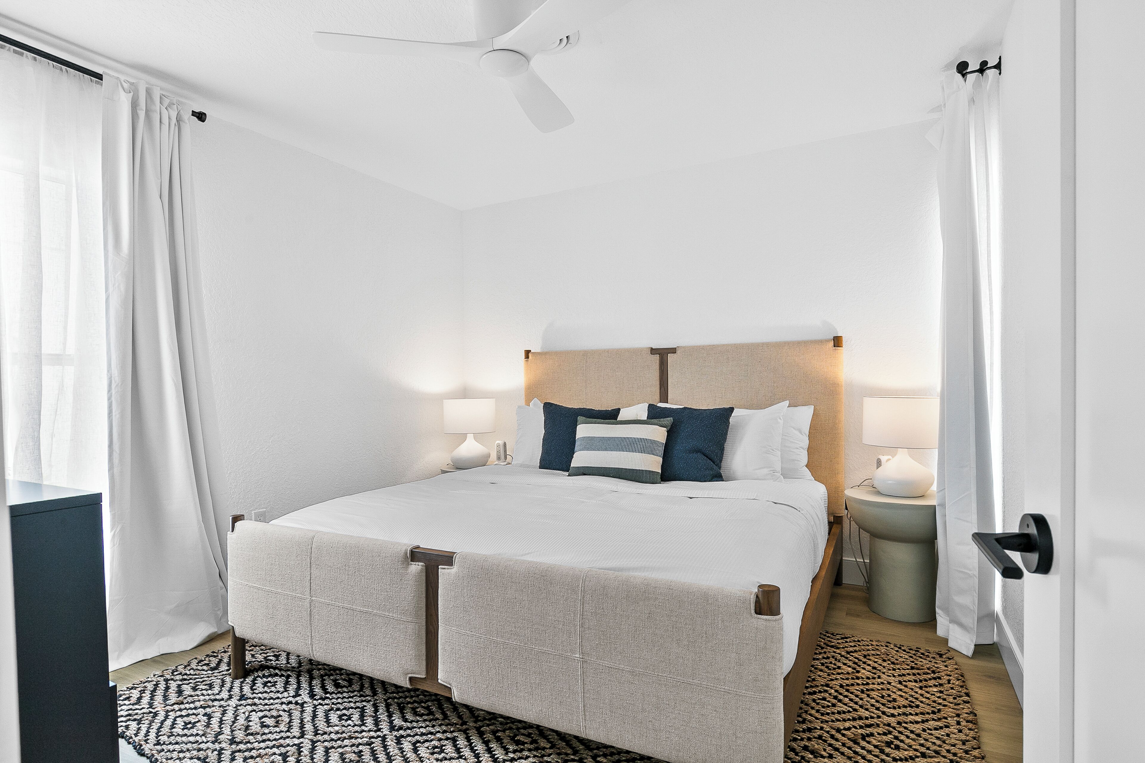 A neatly arranged bedroom with a bed, bedside lamps, a ceiling fan, and a patterned rug.