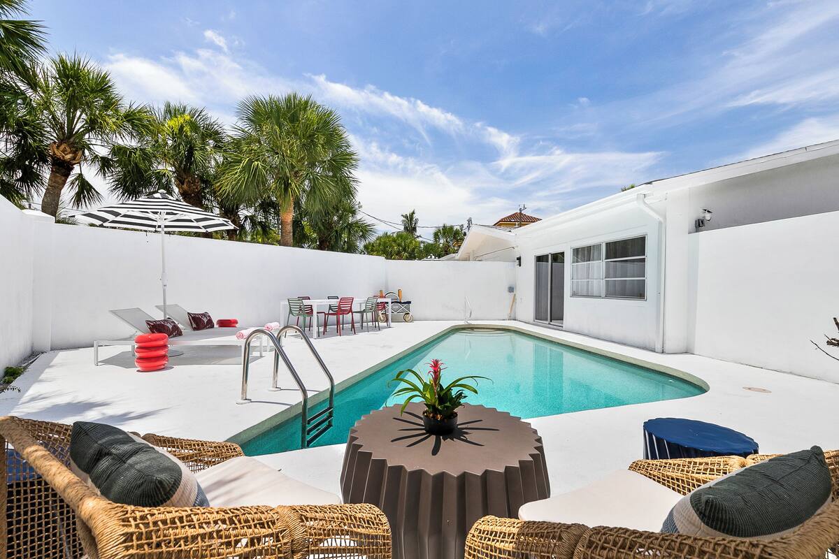 A swimming pool with lounge chairs and a table, surrounded by white walls and palm trees.