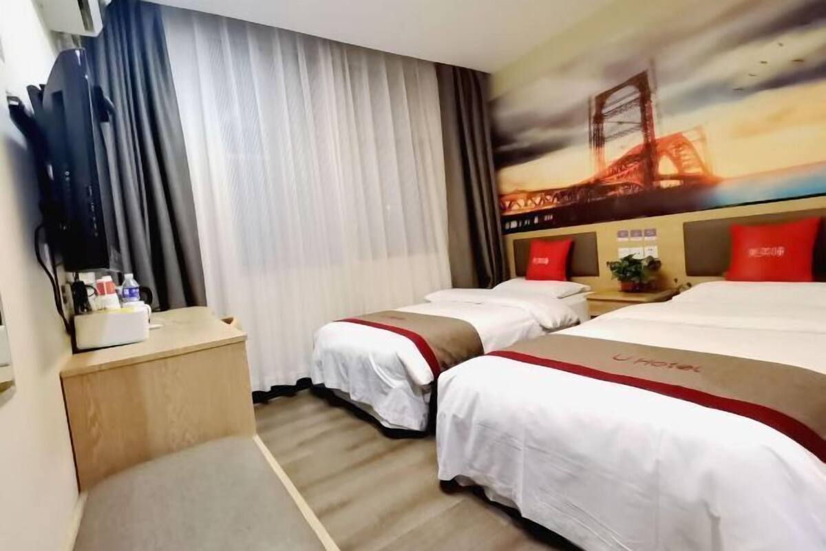 A hotel room with two beds, a TV on a stand, a desk with a lamp, and a painting of a bridge.