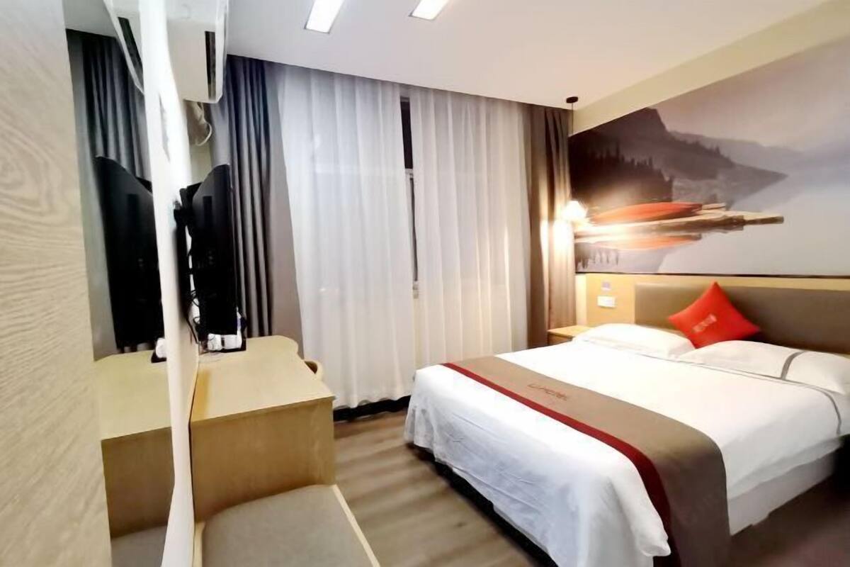 A hotel room with a bed, a desk, a chair, a mirror, a window with curtains, and a wall-mounted artwork.