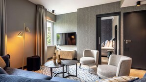 Ein modernes Hotelzimmer mit einer Couch, zwei Sesseln, einem kleinen runden Tisch, einer Stehlampe, einem Fernseher und einer Tür zu einem weiteren Raum.