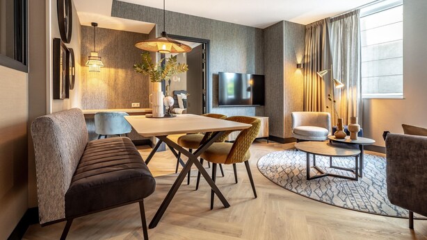 Ein modernes Hotelzimmer mit Essbereich, einer Sitzecke mit rundem Couchtisch und einem großen Fenster mit Vorhängen.