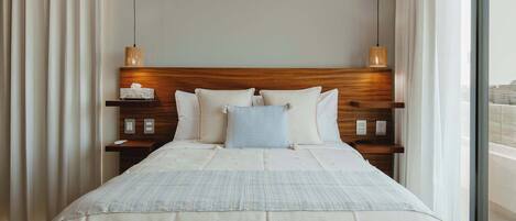 Una cama bien hecha con tablero de madera, dos mesitas de noche con lámparas, y vistas de la ciudad a través de un ventanal grande.