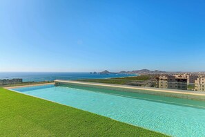 A rectangular swimming pool with clear blue water, surrounded by a well-maintained lawn, and overlooking a coastal cityscape with buildings and a view of the sea. - Arch View Condo Cabo San Lucas • 2nd Floor | Rooftop Pool | 203 (Cabo San Lucas)
