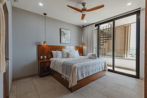 Une chambre à coucher comprenant un lit, une tête de lit en bois, des tables de chevet, un ventilateur de plafond, une fenêtre avec des rideaux et une vue sur un escalier en colimaçon.