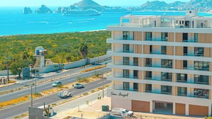A modern hotel with a name sign, located near a coastal road with a view of the sea and rocky formations. - Rooftop Infinity Pool Condo Cabo San Lucas • Modern 2BR El Tezal | 202 (Cabo San Lucas)