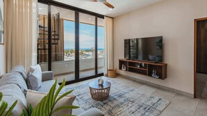 A modern living room with a large TV, a sofa, a coffee table, and a view of the city through floor-to-ceiling windows. - Rooftop Infinity Pool Condo Cabo San Lucas • Modern 2BR El Tezal | 202 (Cabo San Lucas)