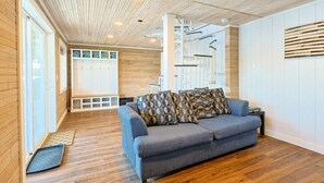 Una sala moderna con un sofá gris, cojines estampados, piso de madera y una escalera al fondo.