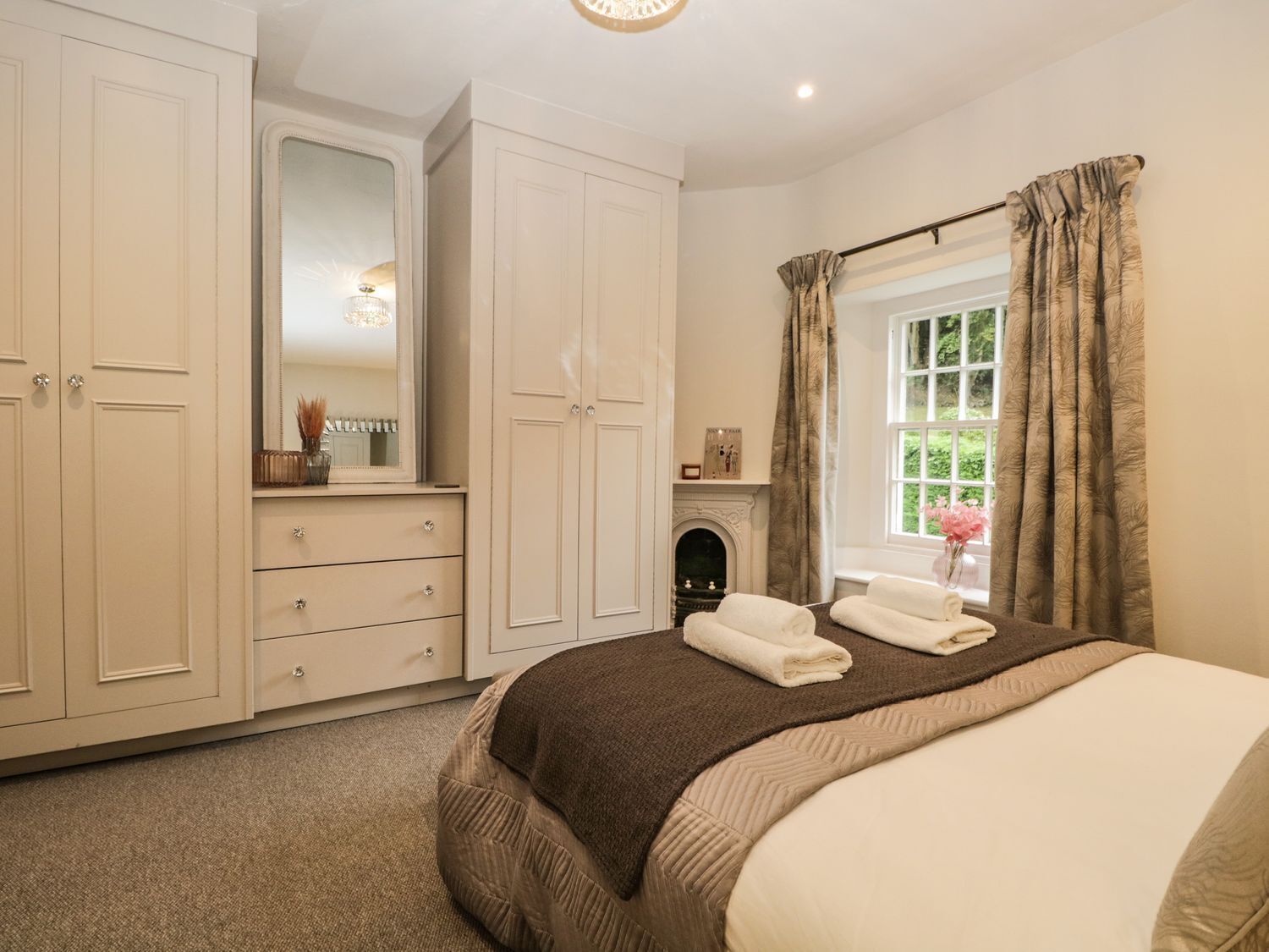 Een slaapkamer met een groot bed, een kaptafel, een spiegel, een open haard en een raam met gordijnen.
