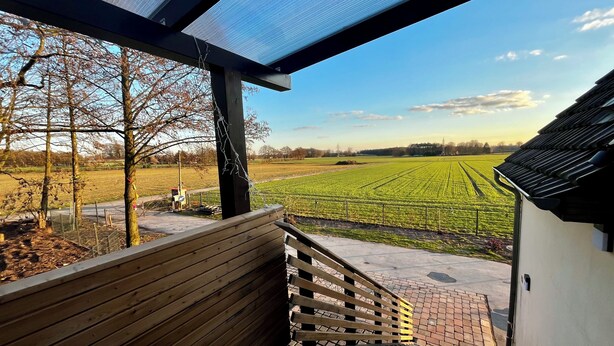 Een overdekt buitengebied met een houten bankje, uitzicht op een groen veld en een gebouw op de achtergrond.