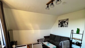 Een moderne loft-achtige woonruimte met een hoge nok, een donkergrijze bank, een houten eettafel en een klein keukengedeelte.