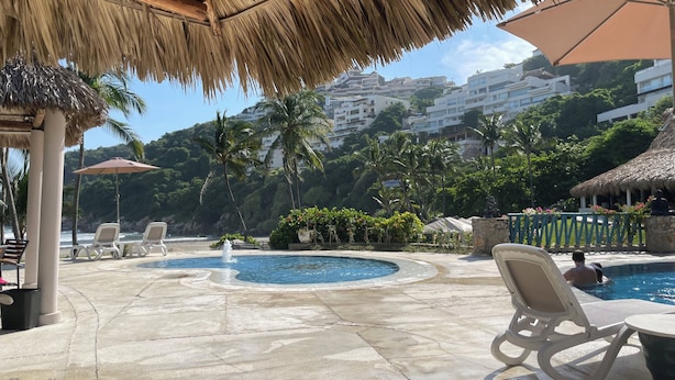 Un espace piscine doté de chaises longues, d’un toit de chaume et offrant une vue sur une colline parsemée de bâtiments.