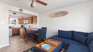 Ein modernes Wohnzimmer mit einem blauen Sofa, einem Holztisch und einem gemusterten Teppich. Im Hintergrund ist eine Küche mit Holzschränken und einer Mikrowelle zu sehen.