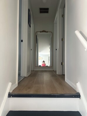 Un couloir doté d’un plancher en bois, de murs blancs et d’une porte au fond, qui donne sur une pièce où se trouve un lit.