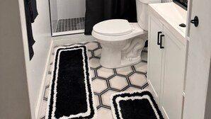 A compact bathroom with a white toilet, a shower area, and a black shower curtain. There are white hexagonal tiles on the floor, and a black and white rug is placed in front of the toilet.