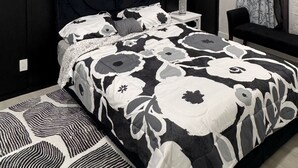 A bedroom with a black headboard, a bed with a black and white floral bedspread, a ceiling fan, a nightstand with a lamp, and a window with floral curtains.