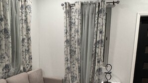 A living room with a beige sectional sofa, floral curtains, a patterned rug, and a white side table with a speaker.