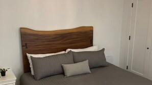 Een slaapkamer met een houten hoofdbord, een groot bed met grijze beddengoed, een nachtkastje met een lamp, en een deur op de achtergrond.
