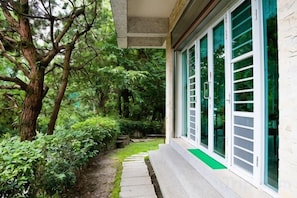 A building with a pathway leading to a door with a green mat, surrounded by lush greenery. - Qingjing Tixiang Panoramic Hostel (Nantou)