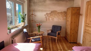 Une pièce confortable avec un lit, une chaise, une petite table sur laquelle est posé un vase de fleurs, un radiateur et une fenêtre agrémentée d’une plante.