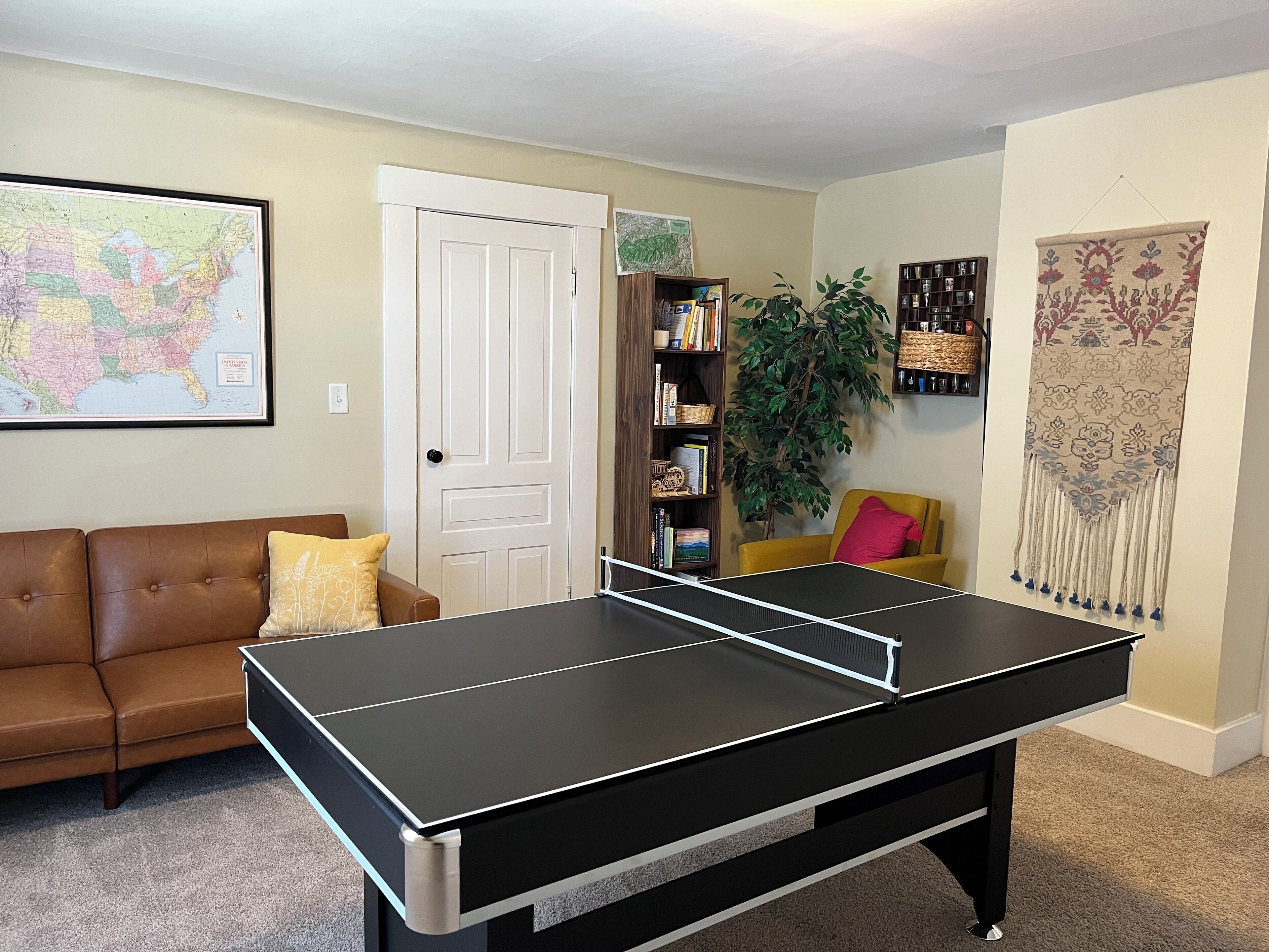 Un salon avec une table de ping-pong, un canapé en cuir marron, une bibliothèque, une plante en pot et une carte accrochée au mur.