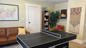 Un salon avec une table de ping-pong, un canapé en cuir marron, une bibliothèque, une plante en pot et une carte accrochée au mur.