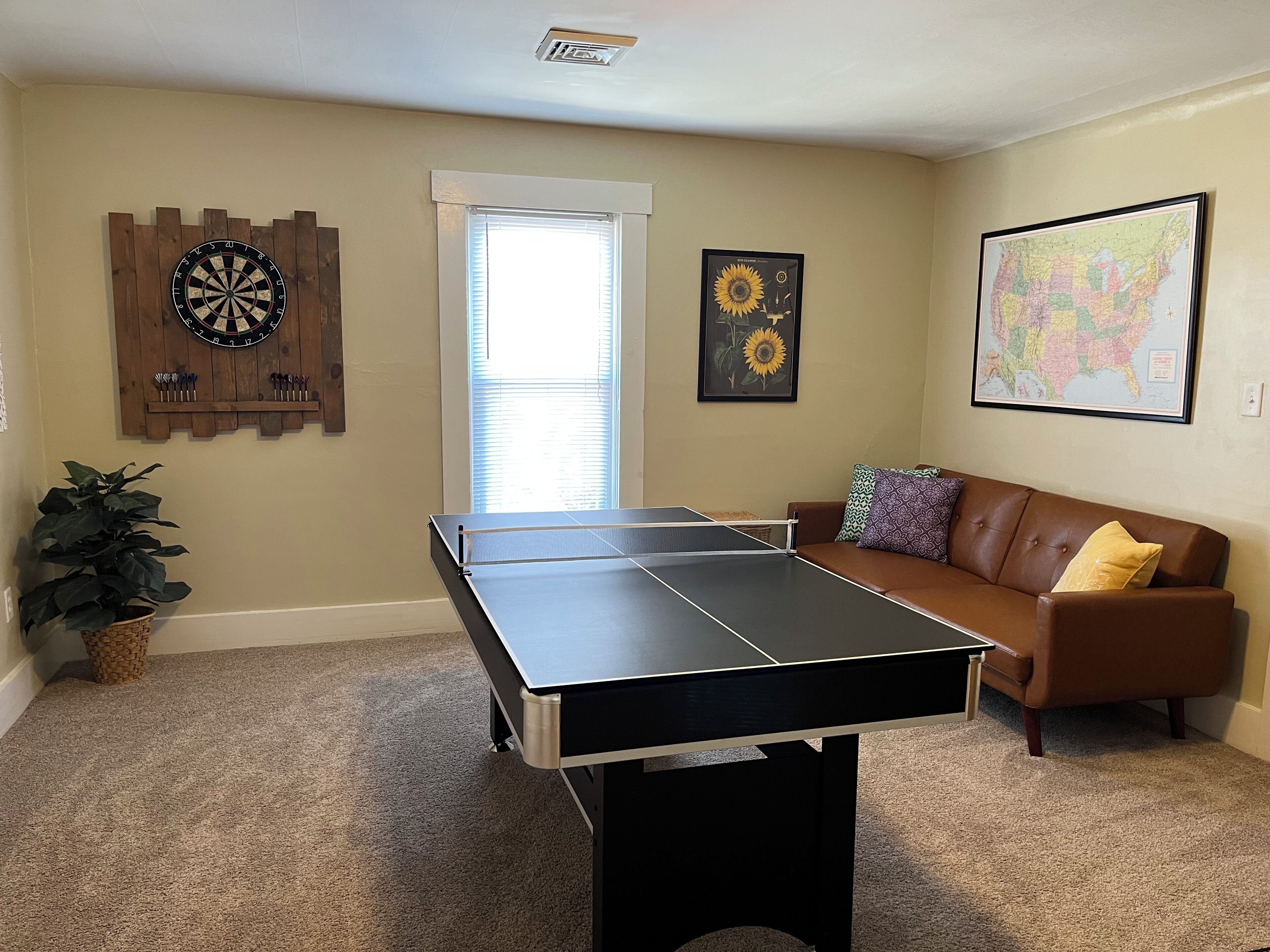 A living room with a ping pong table, a couch, a dartboard, and a potted plant.