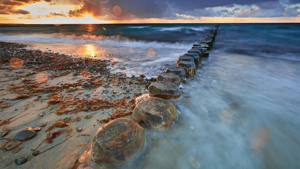 Une plage bordée d’une rangée de rochers couverts d’algues qui s’enfoncent dans l’océan, sous un ciel embrasé par le coucher du soleil.