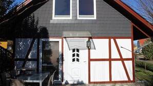Une maison avec un toit en pente, deux fenêtres, une porte et un petit porche comprenant une table et des chaises.