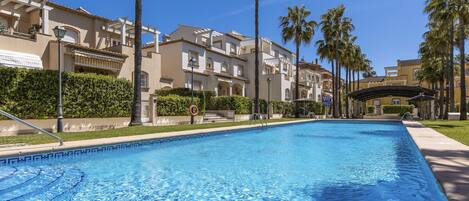 A swimming pool with a clear blue water surface, surrounded by palm trees and a row of beige buildings with balconies.