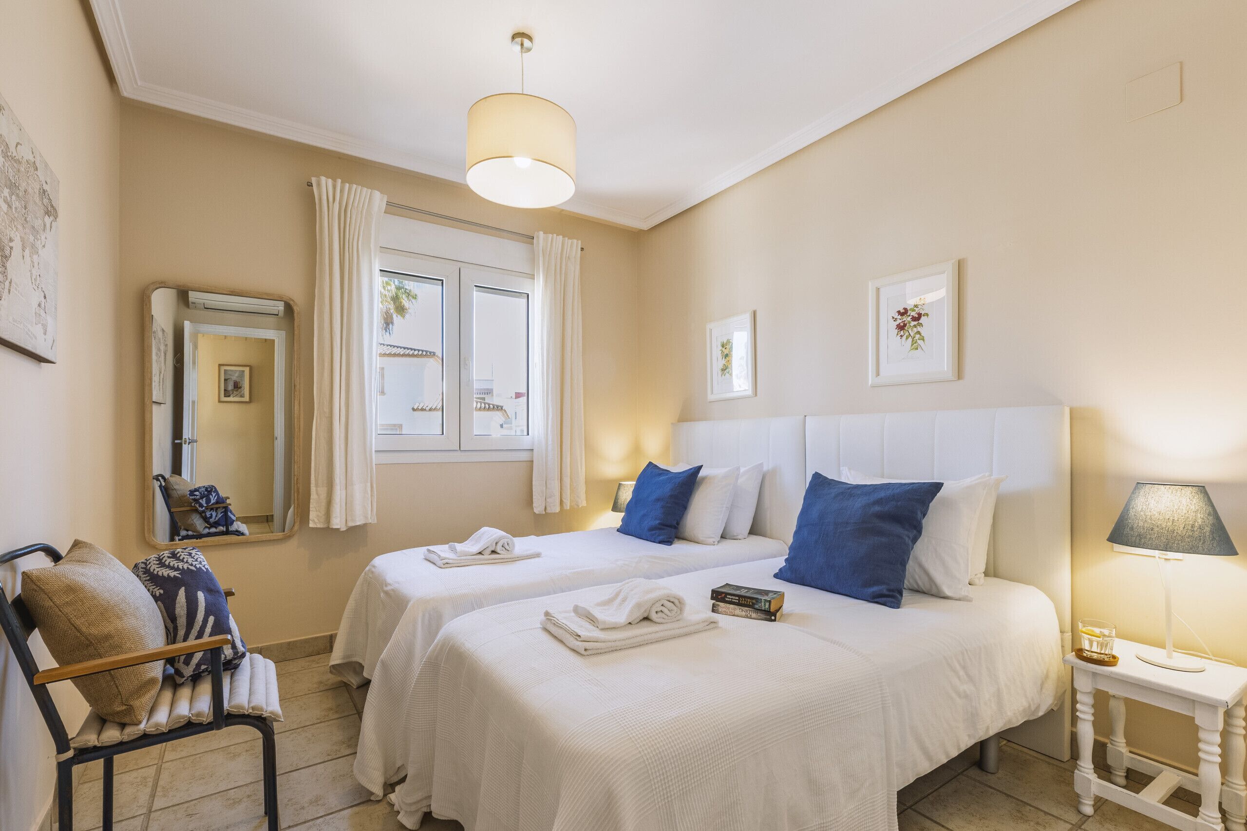 A hotel room with two beds, a mirror, a chair, a bedside table with a lamp, and a window with curtains.