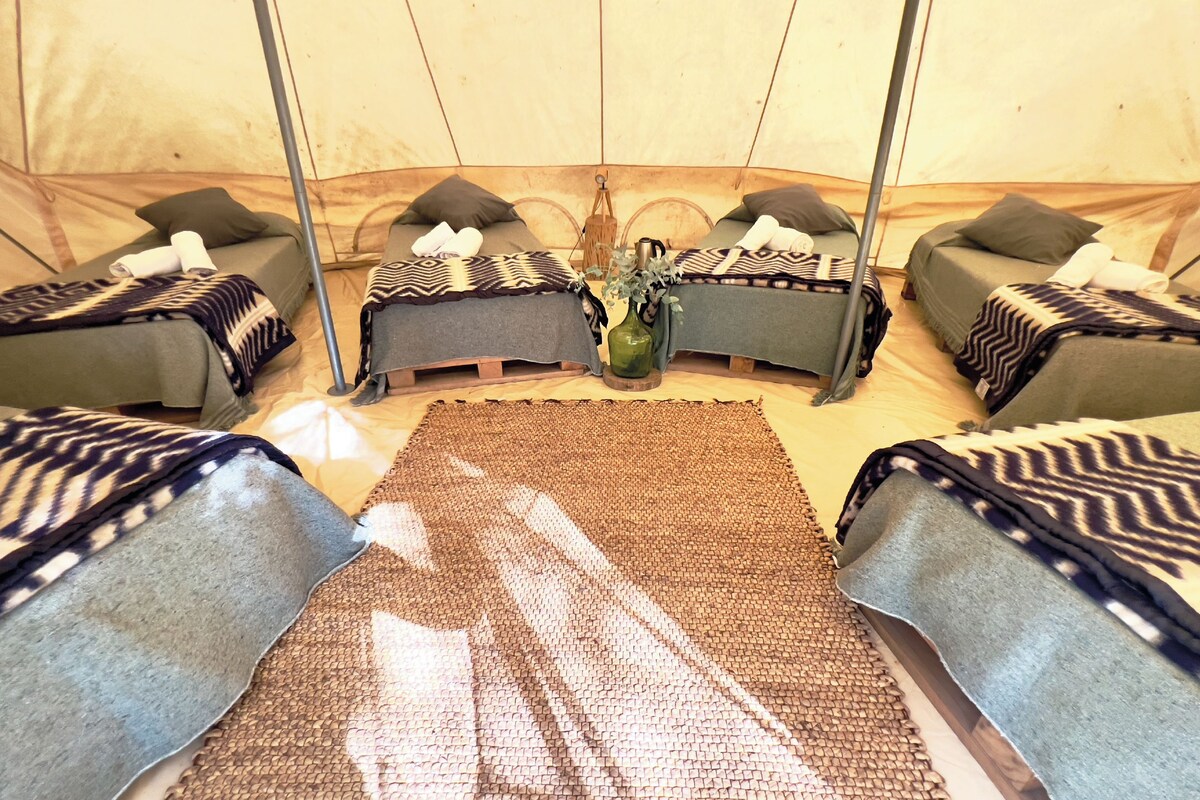 A tent with four beds, each with a patterned blanket and pillows. A central area has a woven mat and a vase with flowers.