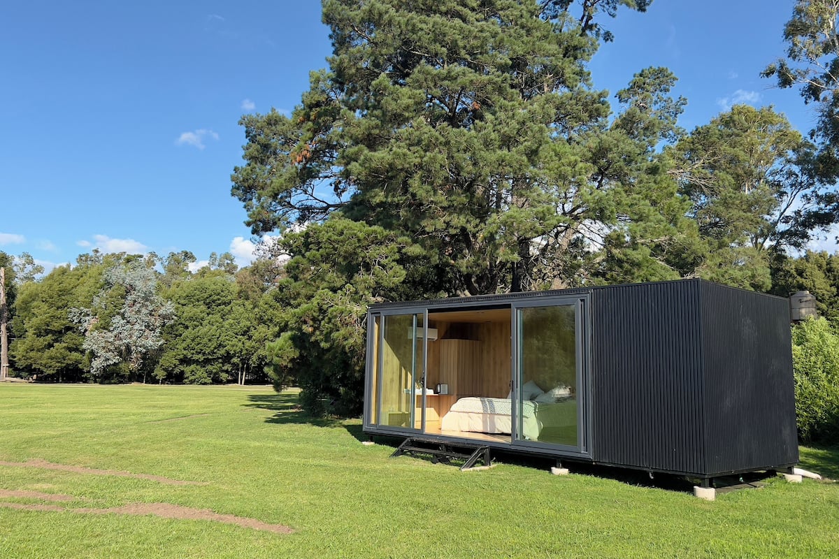 A modern, compact cabin with a glass door, situated in a grassy field with tall trees in the background.