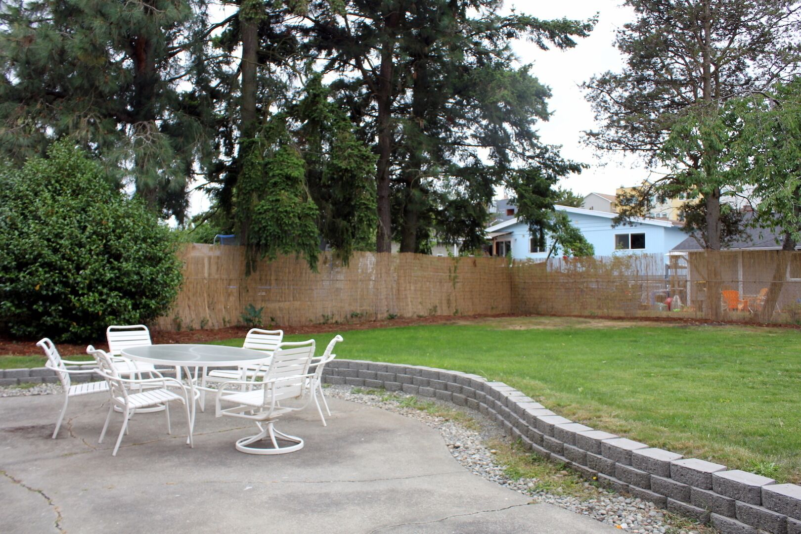 Un jardin avec une table ronde et des chaises, entouré d’une bordure en briques, une clôture en bois et des arbres en arrière-plan.